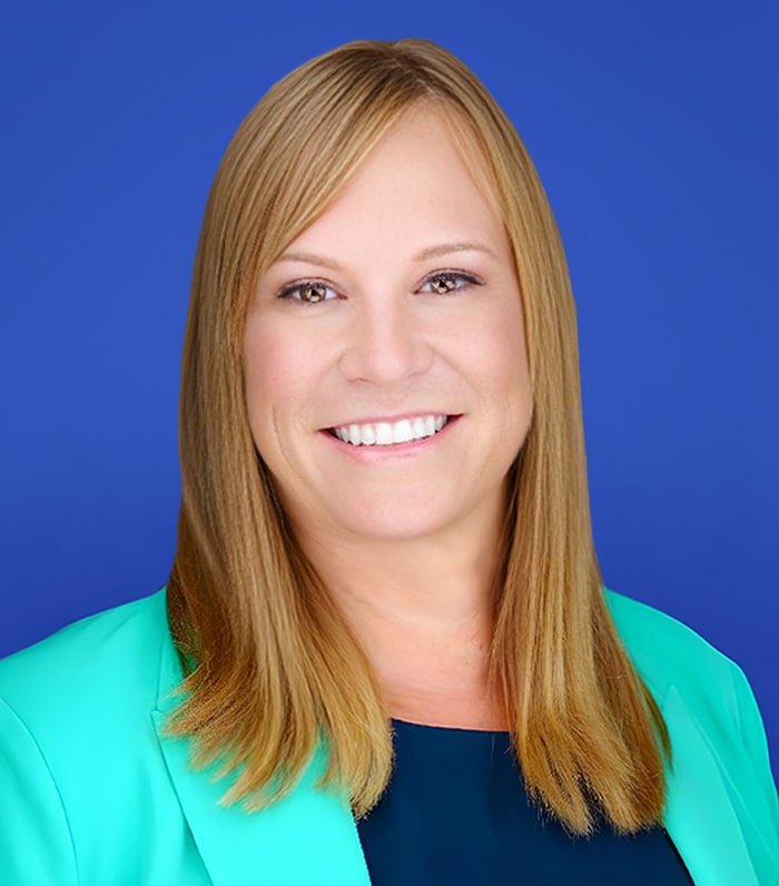 A woman with straight, light brown hair and bangs smiles at the camera. She is wearing a teal blazer over a dark top, with a solid blue background behind her.