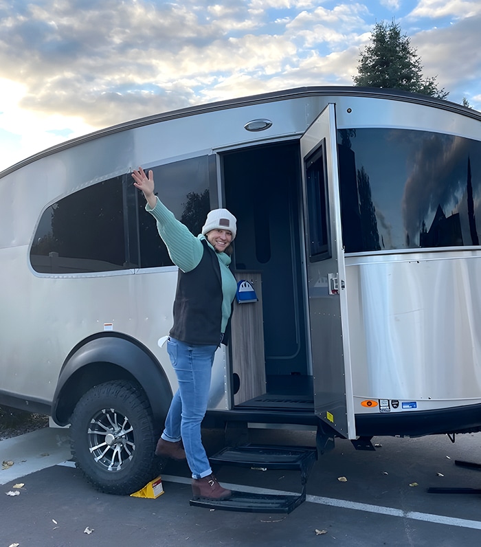 A smiling person in a beanie and vest stands on the steps of a modern silver camper trailer, waving with one hand. The trailer door is open, and the sky is partly cloudy.