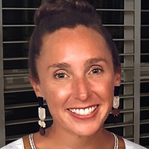 A woman with brown hair in a bun smiles at the camera. She is wearing large, dangling earrings and a white top. Dark blinds or shutters are visible in the background.