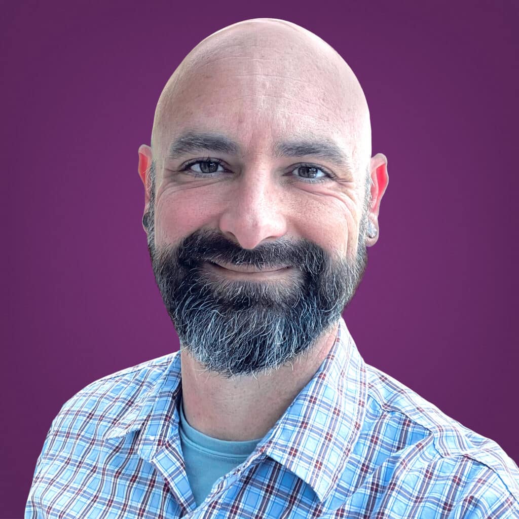 A smiling bearded man with a bald head wearing a plaid shirt and a blue t-shirt, posed in front of a plain purple background.