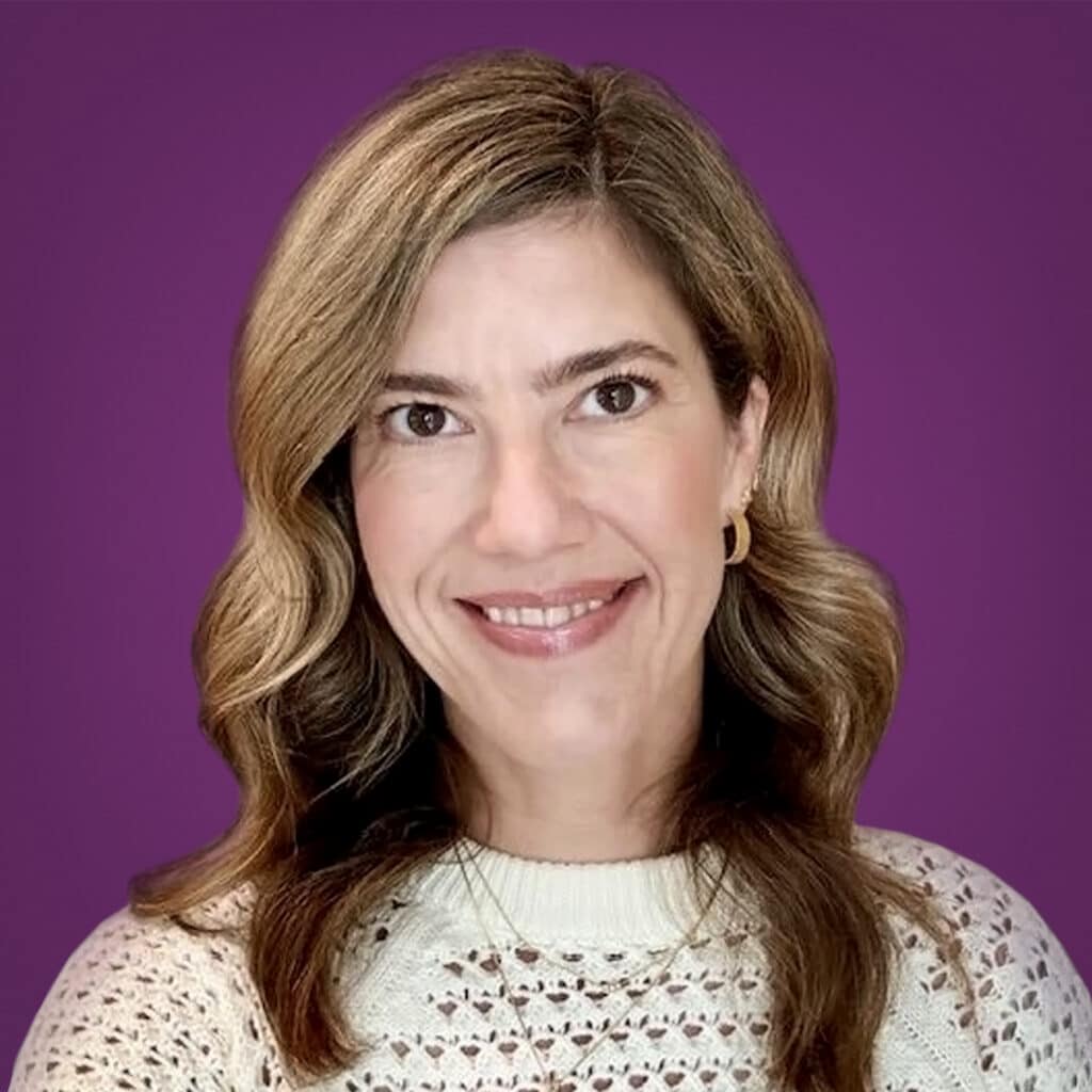 A woman with wavy, shoulder-length blonde hair smiles at the camera. She is wearing a cream-colored knit sweater and gold hoop earrings, with a purple background behind her.