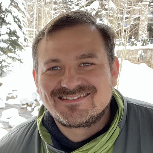 A man with brown hair and a beard is smiling outdoors in a snowy setting, wearing a gray jacket and a green scarf. Snow-covered trees and stone structures are visible in the background.