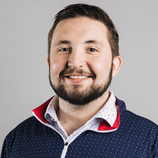 A man with short brown hair and a beard smiles at the camera. He is wearing a navy blue zip-up jacket with white polka dots over a white shirt with a red and blue pattern. The background is light gray.