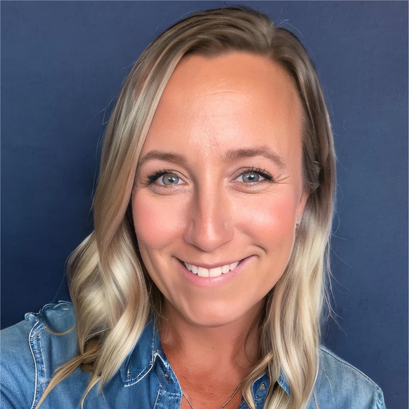 A woman with long, blonde wavy hair smiles at the camera. She is wearing a blue denim shirt and is posed in front of a solid dark blue background.