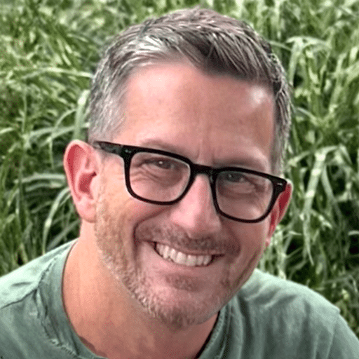 A smiling middle-aged man with short gray hair, wearing black-rimmed glasses and a green shirt, is pictured outdoors with green foliage in the background.