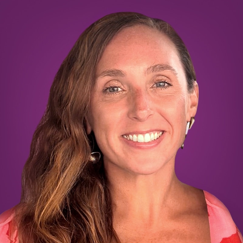 Smiling woman with long wavy brown hair wearing a pink top and gold earrings, posing confidently in front of a solid purple background, as if ready to present a benefits administration software demo.
