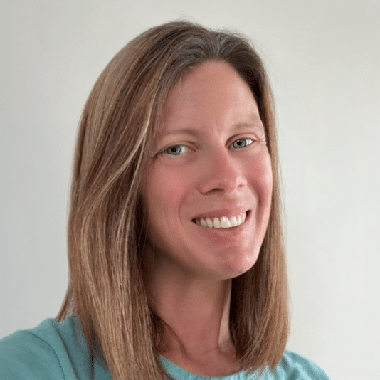 A woman with straight, shoulder-length light brown hair smiles at the camera. She is wearing a light blue top and is posed against a plain, light-colored background.