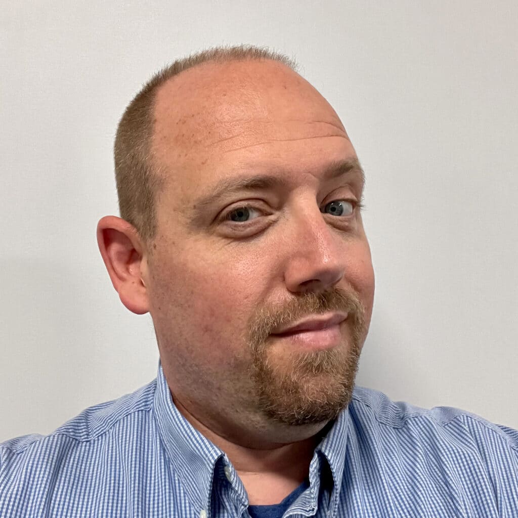 A man with short light brown hair, blue eyes, and a trimmed goatee wearing a blue striped shirt looks at the camera with a slight smile, embodying confidence in the face of HR leadership challenges, against a plain white background.