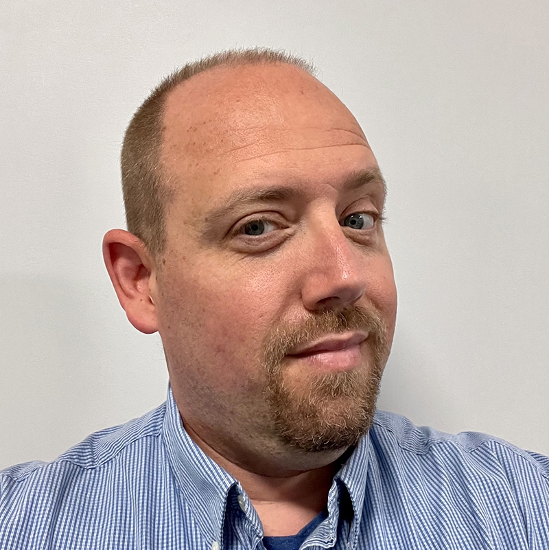 A man with short light brown hair and a goatee, wearing a light blue striped button-up shirt, looks at the camera with a neutral expression against a plain white background—capturing the thoughtful demeanor often seen in professionals navigating HR leadership challenges.