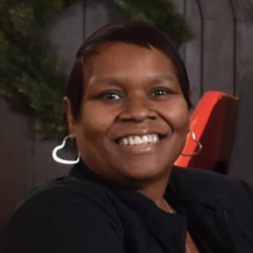 A smiling person with short hair, wearing large hoop earrings and a dark top, sits in front of a blurred background with greenery—embodying confidence and approachability often needed to navigate HR leadership challenges.