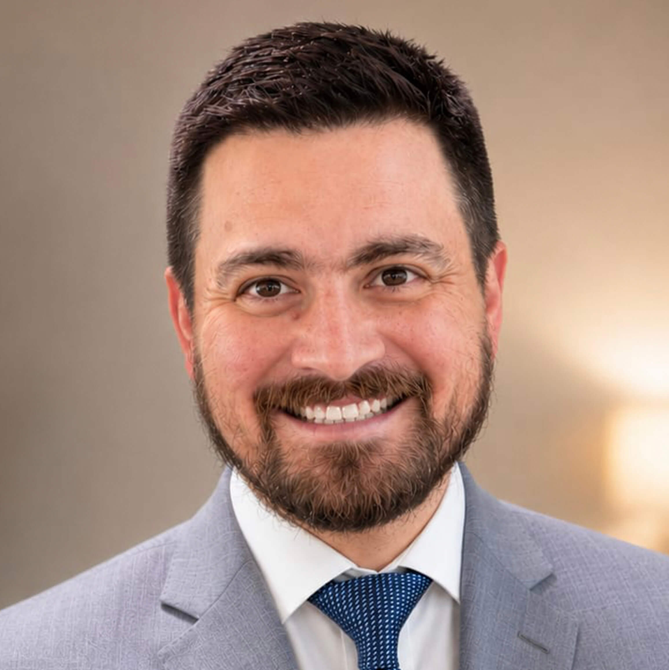 A man with short dark hair and a beard, wearing a light gray suit, white shirt, and blue tie, smiles in front of a blurred, neutral background—exuding confidence and approachability amid today’s HR leadership challenges.