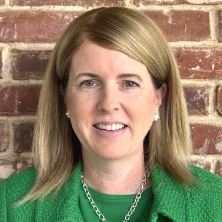 A woman with straight, shoulder-length blonde hair is smiling in front of a brick wall. She is wearing a green jacket, a green top, a silver necklace, and pearl earrings.